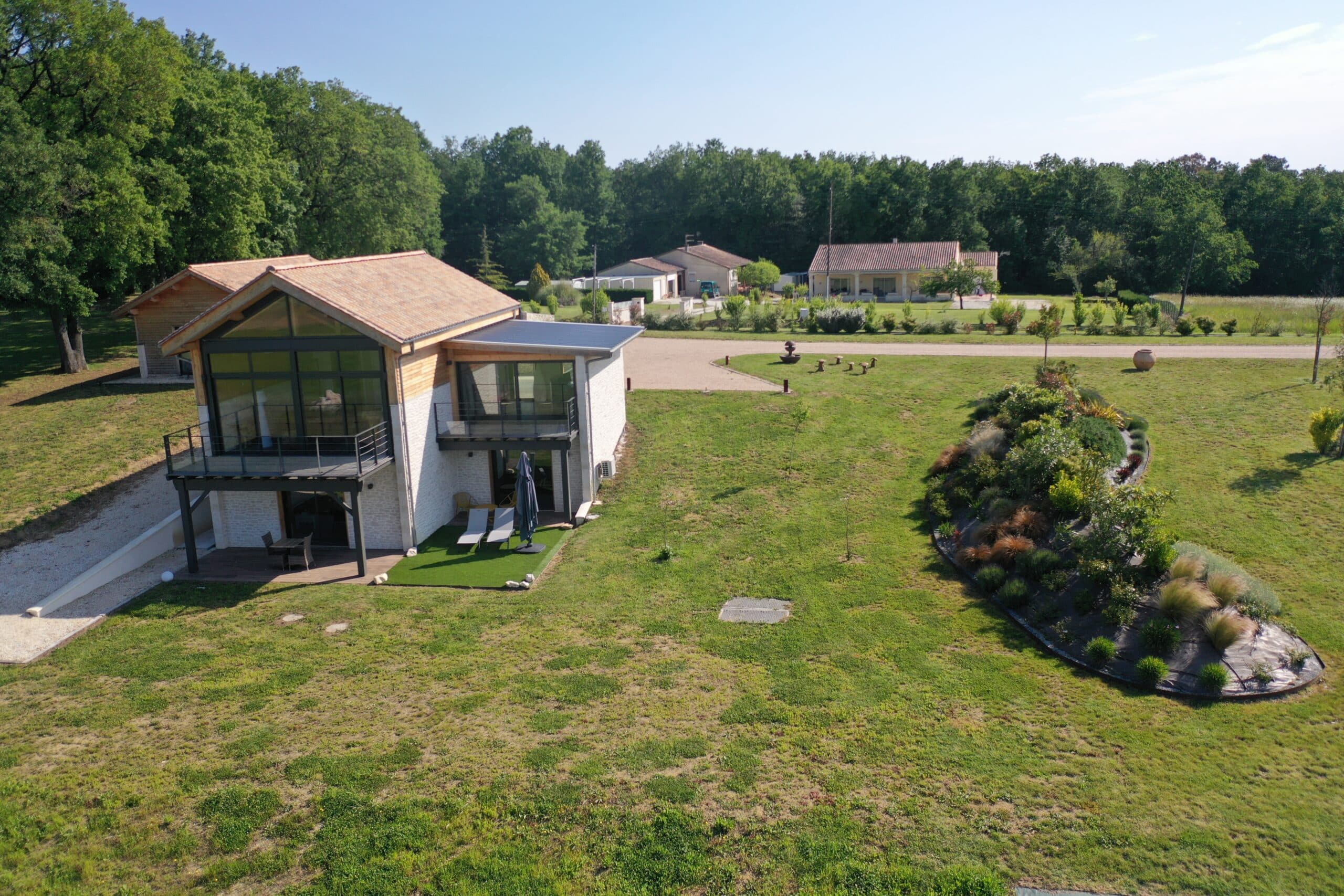 Maison moderne avec façade vitrée et terrasses, sur grande pelouse arborée. Environnement paisible avec autres maisons et jardin paysager.