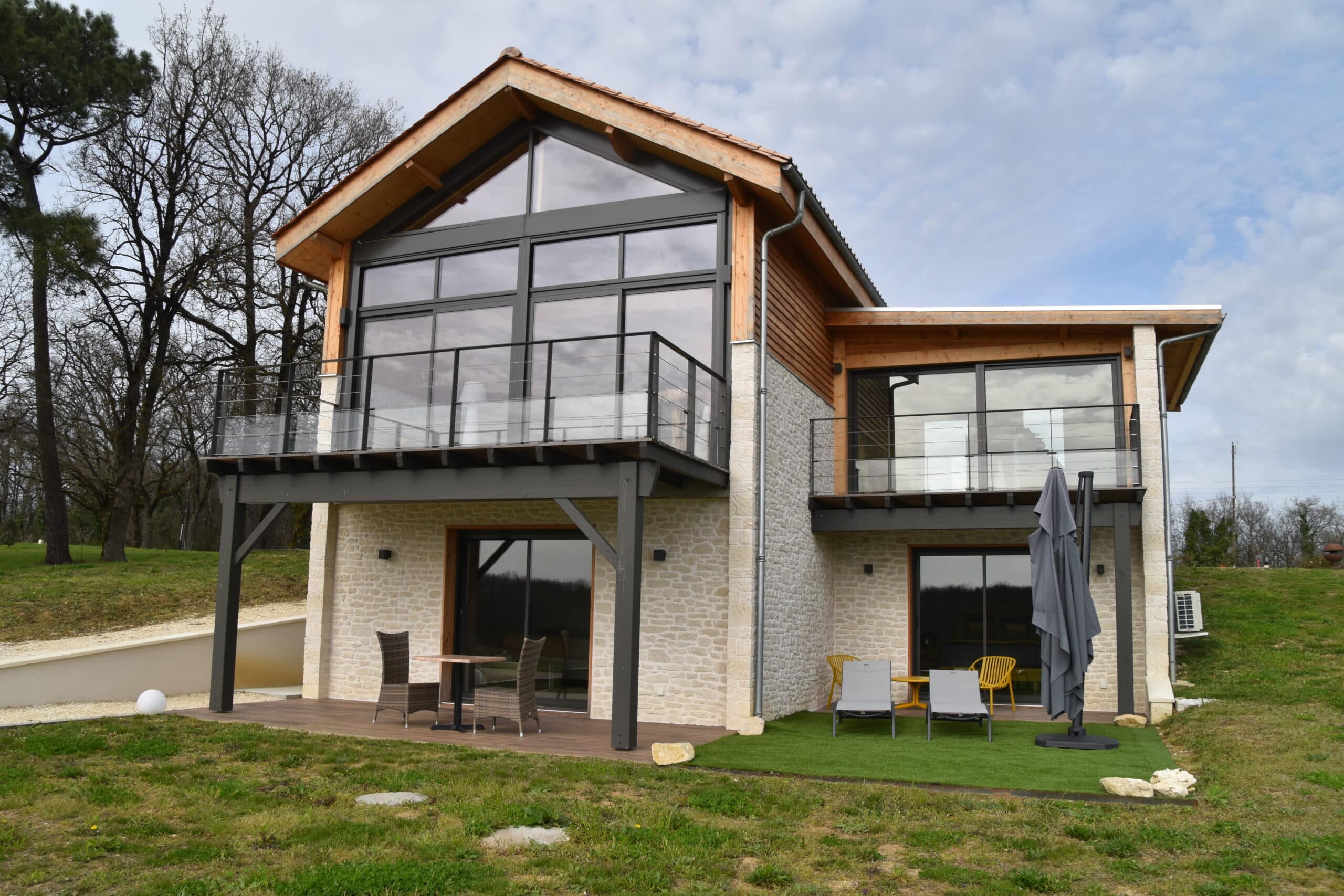 Maison moderne en pierre et bois avec de grandes baies vitrées, balcons aménagés, terrasses en bois et pelouse verte.