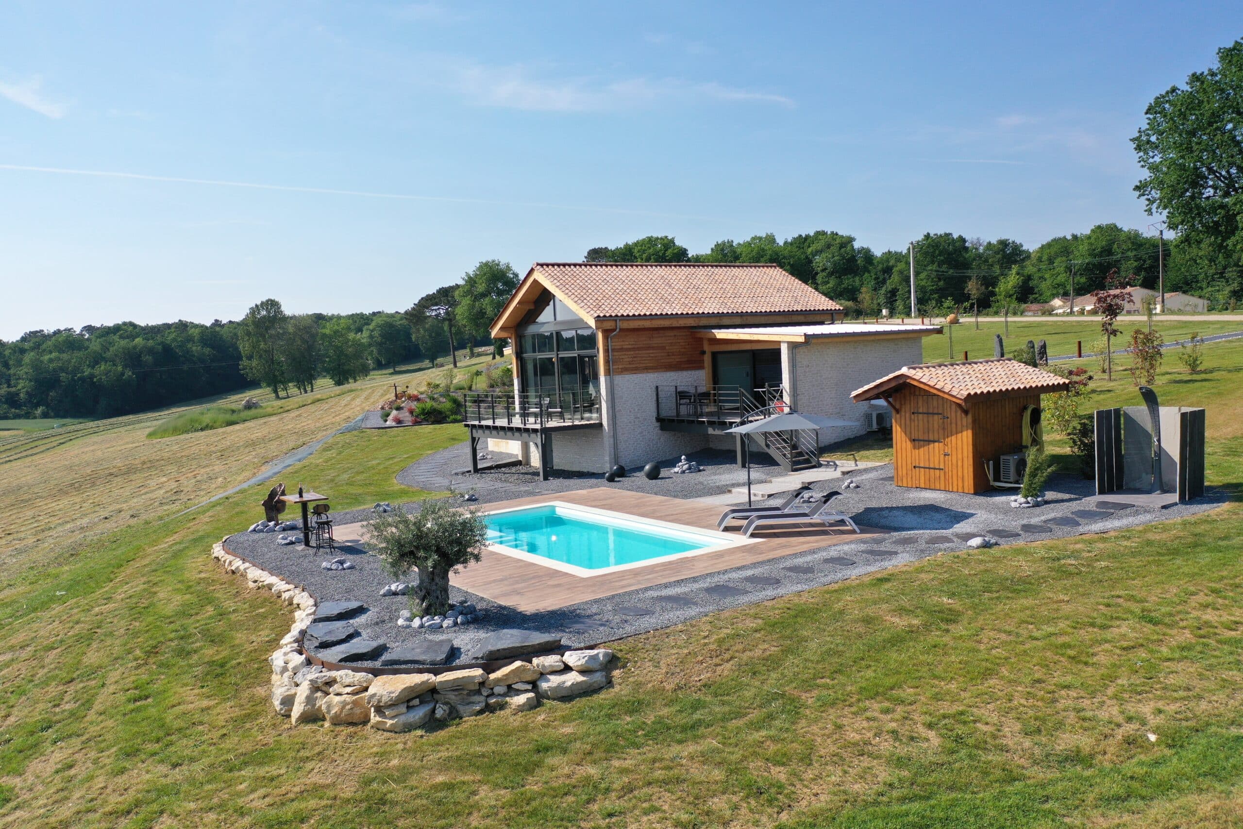 Villa moderne avec piscine, terrasse bois, transats, parasol et douche extérieure. Jardin gravier, champs et forêts sous ciel bleu.