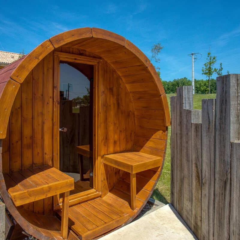 Sauna tonneau en bois verni avec porte vitrée et deux bancs intérieurs, dans un jardin verdoyant sous un ciel bleu.