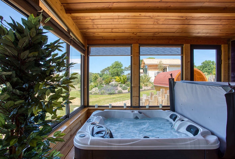 Jacuzzi bouillonnant dans une véranda en bois avec vue sur un jardin verdoyant et des structures modernes sous un ciel bleu.
