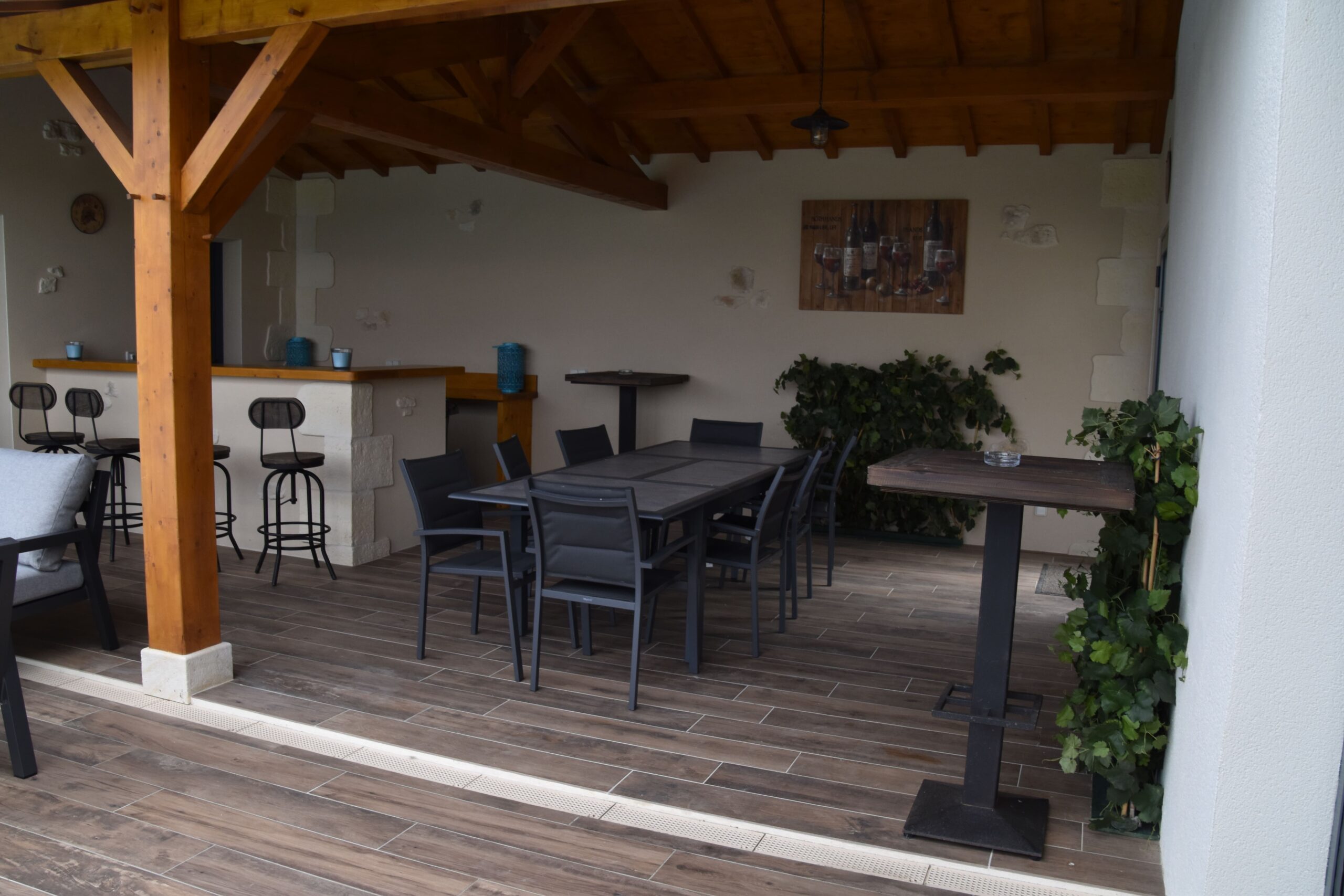 Terrasse couverte extérieure : bar, tabourets, grande table à manger, chaises. Structure bois, sol carrelage aspect bois. Décoration tableau vins et plantes.