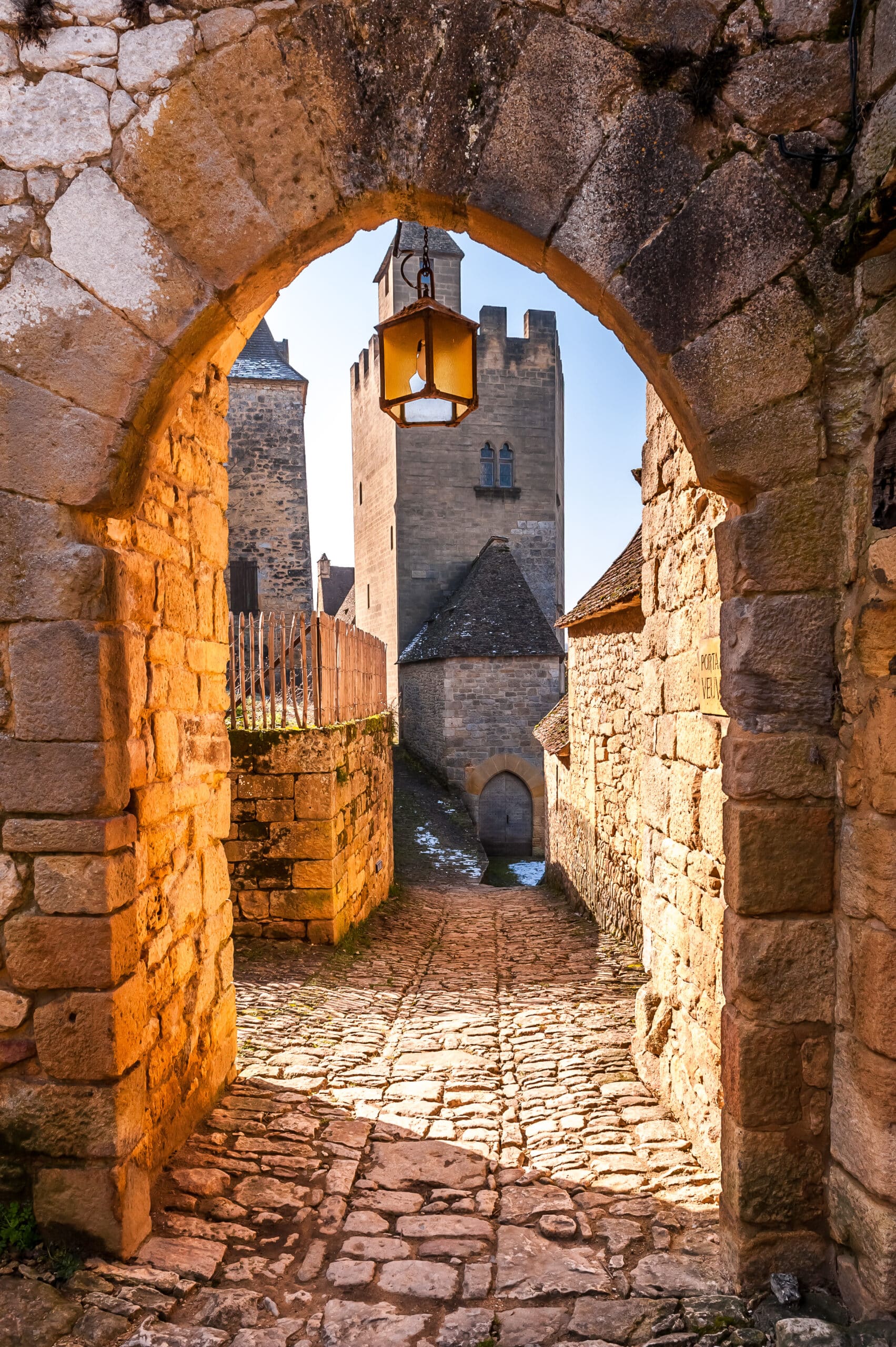 Chemin pavé sinueux sous une ancienne arche en pierre, lanterne suspendue. Tours médiévales et inscription "PORTA VE" au loin.