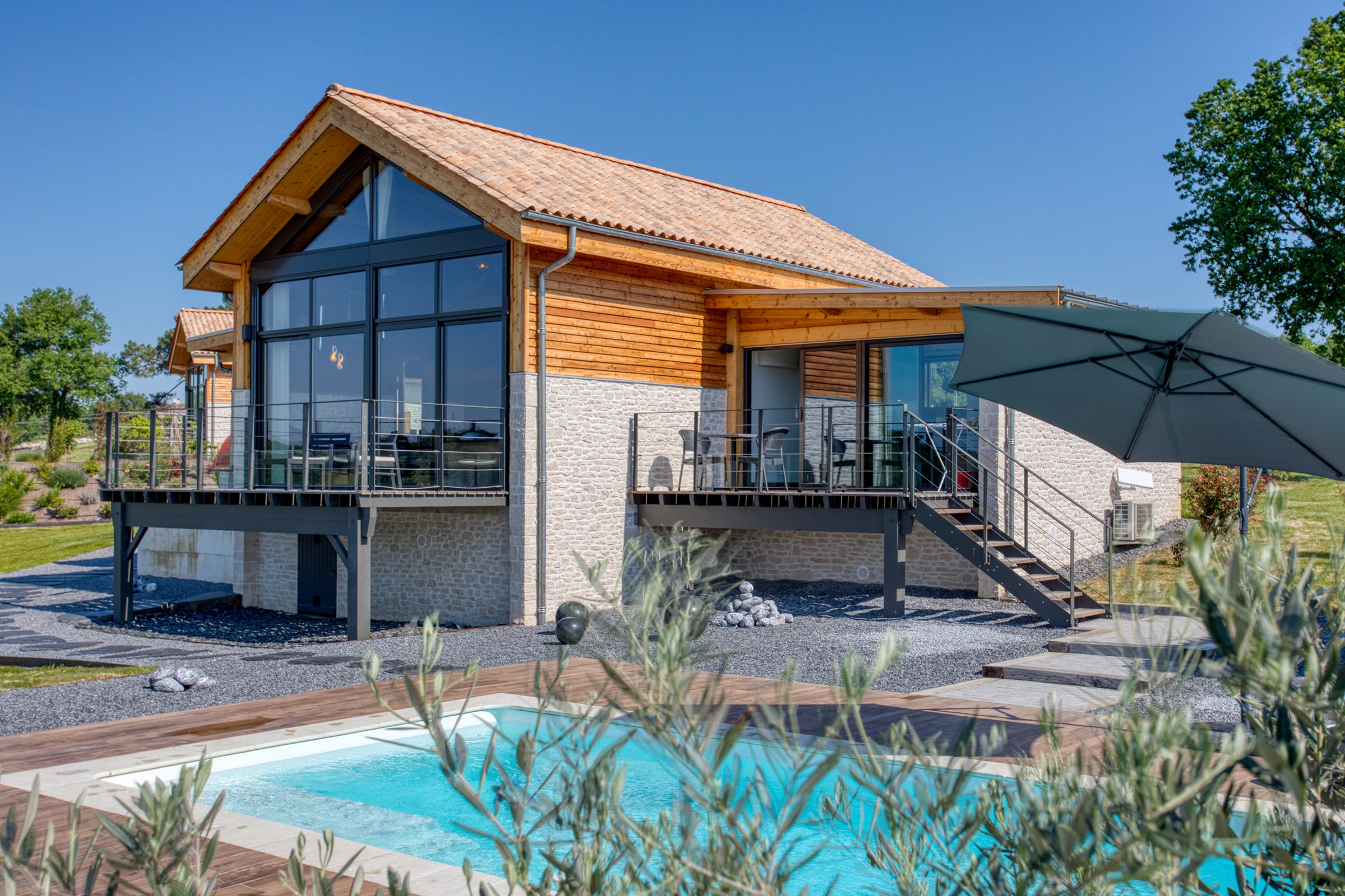 Maison moderne avec façade bois et pierre, grande baie vitrée, terrasse avec balustrade, piscine extérieure, sous ciel bleu.