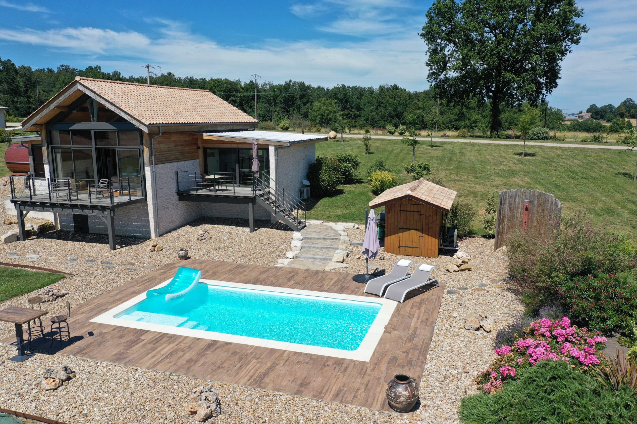 Maison moderne avec toits tuile/métal, grandes baies vitrées, piscine bleue, terrasse en bois, chaises longues, douche extérieure et chalet jardin. Ciel bleu.
